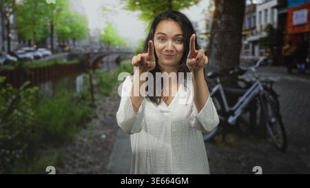 Frau zeigt beide Zeigefinger auf die Kamera auf der Kanalstraße in der Nähe eines geparkten Fahrrads in weißer Bluse; verspieltes Selbstvertrauen. Stockfoto