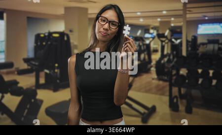 Thailändische Frau hält in einem Fitnessstudio ein weißes Puzzleteil neben ihr Gesicht, trägt eine Brille und ein ärmelloses schwarzes Oberteil mit lässigem Armband; Neugier auf Nachdenklichkeit. Stockfoto