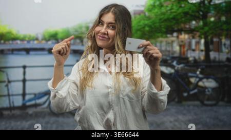 Frau, die eine Kreditkarte hält, zeigt auf der Straße an einer Kanalbrücke mit dem Finger auf die Karte; Selbstvertrauen stärkt ein Lächeln. Stockfoto