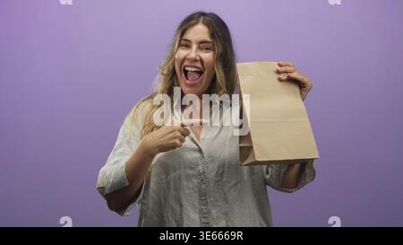 Frau zeigt mit dem Finger auf die Papiertüte im Studio, während sie lächelt, Mund und Zähne sichtbar; fröhliche Enthüllung. Stockfoto