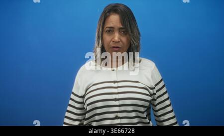 hispanische Frau mittleren Alters in gestreifter Strickjacke, neutrales Gesicht mit geschlossenen Augen und erhobenen Armen vor blauem Studio-Hintergrund; ruhige Besinnung Stockfoto