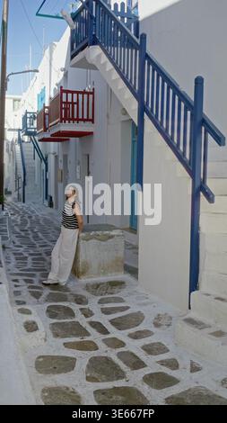 Frau, die auf einer Steinstraße in mykonos in der Nähe von weißen Gebäuden mit blauen Treppen und roten Balkonen läuft; Ruhe. Stockfoto