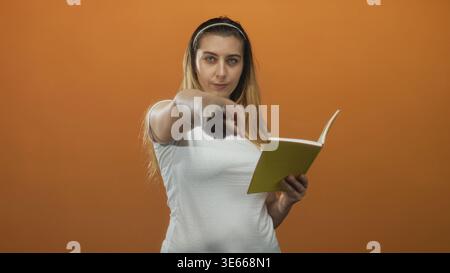 Die Frau zeigt mit der ausgestreckten Hand das grüne Notizbuch in der Hand und trägt ein weißes T-Shirt vor orangefarbenem Studio-Hintergrund Stockfoto