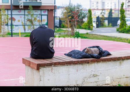 Person im Hoodie sitzt auf der Bank neben Sleeping Cat on Jacket am Urban Park Court Stockfoto
