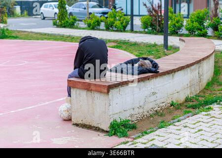 Person im Hoodie sitzt auf der Bank neben Sleeping Cat on Jacket am Urban Park Court Stockfoto