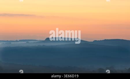 Neblige Hügel, die sich bei Sonnenaufgang oder Abenddämmerung unter einem warmen, abgestuften Himmel bilden, schaffen eine ruhige und stimmungsvolle natürliche Landschaft. Stockfoto