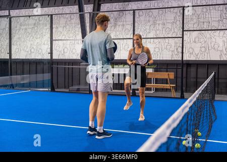 Trainingspartner, die Schläger halten, die Padel-Ball über dem Netz auf dem blauen Platz mit Bank und Zaun schlagen Stockfoto