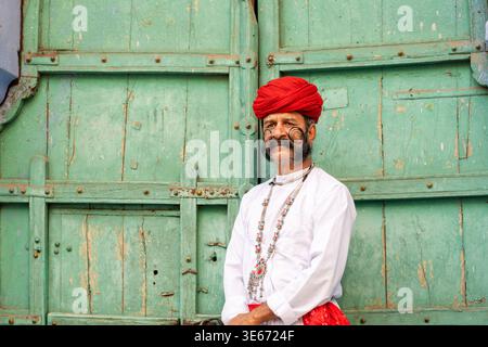 Porträt eines Indianers in der Blauen Stadt Jodhpur, Rajasthan. Er hat einen langen, lockigen Schnurrbart mit einem roten Turban, der vor grünen Türen steht. Stockfoto