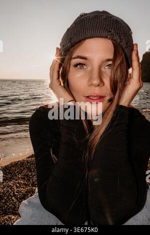 Frau, Porträt, Sonnenuntergang. Junge Frau posiert in Strickmütze am Strand zur goldenen Stunde mit dramatischer Beleuchtung. Stockfoto