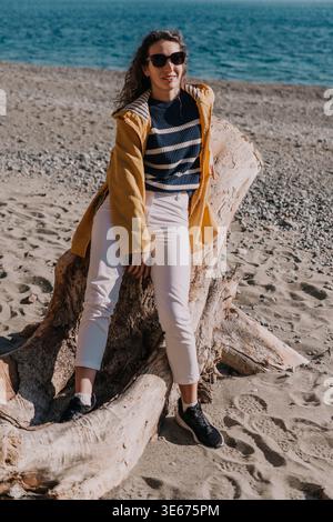 Frau Strand Treibholz sitzt auf einem großen verwitterten Baumstamm und trägt einen gelben Regenmantel am Meer Stockfoto