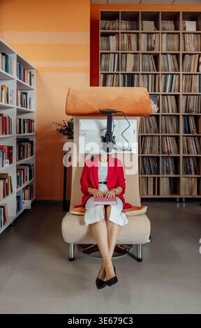 Unerkennbare Frau sitzt in einem modernen Coworking-Raum. Sie trägt einen roten Blazer und ein weißes Kleid und schreibt auf einem Laptop. Bücherregale sind im Hintergrund. Stockfoto