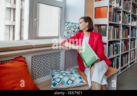 Frau mit kurzen braunen Haaren sitzt auf einer Couch in einer Bibliothek, mit Buch. Bücherregale mit Büchern sind im Hintergrund sichtbar. Stockfoto