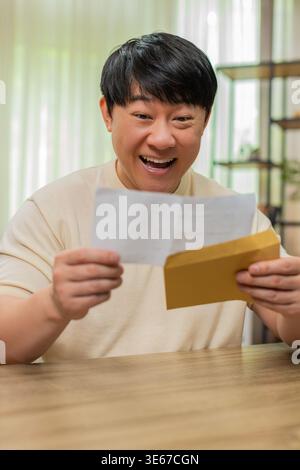 Ein asiatischer Mann, der zu Hause sitzt, liest einen offenen Brief, die Augen erhellen sich, lächelt, als ihn die gute Nachricht erreicht. Der chinesische Kerl faltet sanft Papier, erleichtert und glücklich, genießt positive Worte, die heute warm empfangen werden. Stockfoto