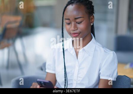 Junge Schwarze Frau, die ihr Smartphone benutzt, während sie drinnen auf einem Sofa sitzt und leise lächelt in einem hellen, modernen Raum. Mobile Kommunikation, Online-Browsing und Stockfoto
