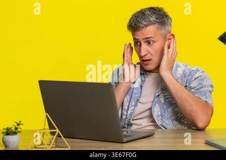 Ein Mann mittleren Alters liest unerwartete E-Mails auf einem Laptop am Tisch, zeigt einen Alarmfokus und lehnt sich dann plötzlich mit weit geöffneten Augen geschockt zurück. Junger Mann, der emotional auf Überraschung reagiert, auf gelbem Hintergrund. Stockfoto