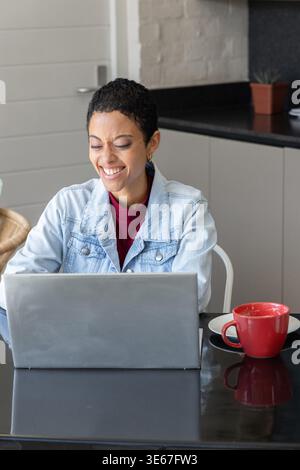 Afroamerikanerin lächelt, in Jeansjacke, arbeitet am silbernen Laptop am Küchentisch mit Becher Stockfoto