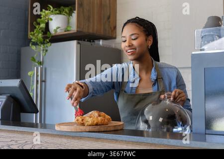Afroamerikanerin in grüner Schürze hinter dem Kaffeekocher lächelnd und mit einer roten Zange zu Gebäck Stockfoto