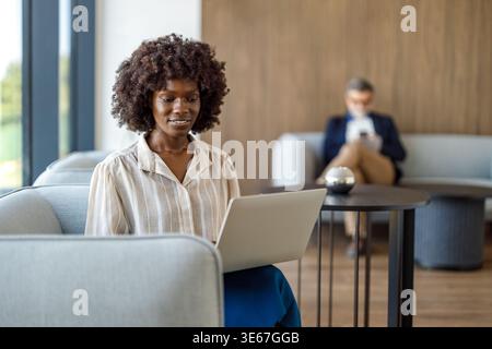 Fokussierte Profi-Frau mit Afro-Frisur, die Daten analysiert, während sie sich auf dem Sofa in einem modernen Büro entspannt Stockfoto