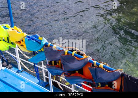 Bunte Schwimmwesten in Gelb, Blau, Orange, Grau und Schwarz hängen an einem blauen Geländer an einem Boot. Das Meer ist im Hintergrund sichtbar, was saf betont Stockfoto