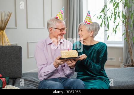 Wünschen Sie sich etwas. Familie Seniorenpaar in Partymütze feiert gemeinsam Geburtstag zu Hause. Ein alter Mann, der Kerzen auf der Geburtstagskuche brennt. Die alte Frau wünscht dem Mann alles Gute zum Geburtstag Stockfoto