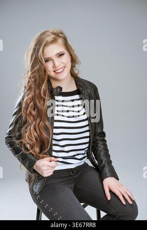 Frau mit langen braunen Haaren sitzt auf dem Stuhl und lächelt. Sie trägt ein schwarz-weiß gestreiftes Hemd und eine Lederjacke. Concept Concept Confidence und Style, während die Frau für ein Foto posiert Stockfoto