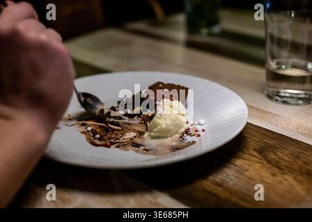 Nahaufnahme einer Hand, die einen Löffel an ein halb gegessen Eis-Brownie-Dessert hält, geschmolzene Schokolade auf einem Teller Stockfoto