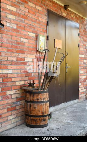 Gartenwerkzeuge, die in Holzfässern neben einer Metalltür gelagert werden, verschiedene Werkzeuge, die herausragen, bereit für den Einsatz im Freien wie Garten oder Landwirtschaft. vertica Stockfoto