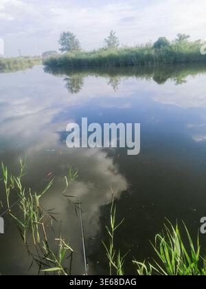 Vertikaler Foto Fluss umgeben von grüner Vegetation fließt ruhig unter hellem Himmel. Reflexion verbindet Himmel und Wasser. Immer noch liegt der Fluss zwischen grünen s Stockfoto