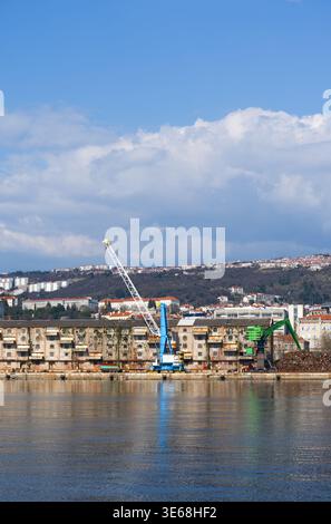 Eine große Aufnahme eines Frachthafens mit schweren blauen und grünen Hafenkranen vor verwitterten Industrielagern. Stockfoto