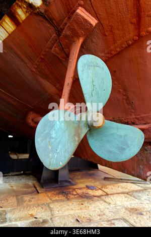 Detaillierte Ansicht eines verwitterten Bronze-Bootspropellers, bedeckt mit türkisfarbener Oxidation, montiert auf einem korrodierten Metallrumpf. Industrieller maritimer Hintergrund Stockfoto