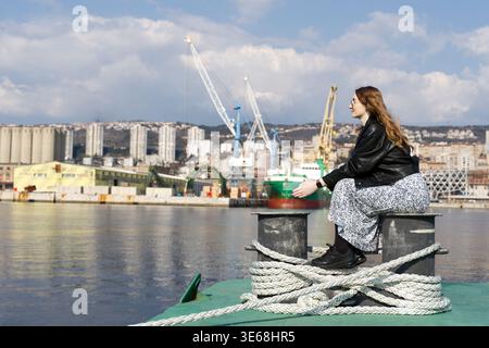Junge Frau in lässigem Outfit, die auf einem Schiffspoller mit Seehafen-Hintergrund sitzt. Weibliche Reisende genießen den Blick auf den Industriehafen und den Frachtschiffen Stockfoto