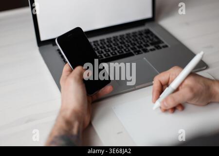 Professioneller männlicher Designer sitzt am Schreibtisch mit Laptop, Tablet und hält Smartphone Stockfoto