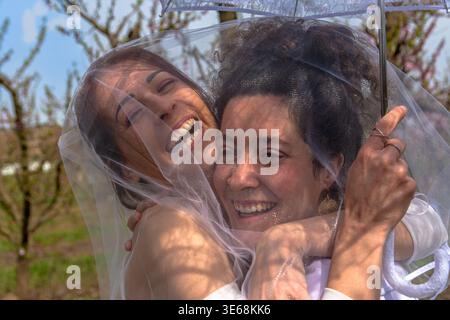 Glückliche Frauen, die sich unter Hochzeitsschleier im Obstgarten umarmen Stockfoto