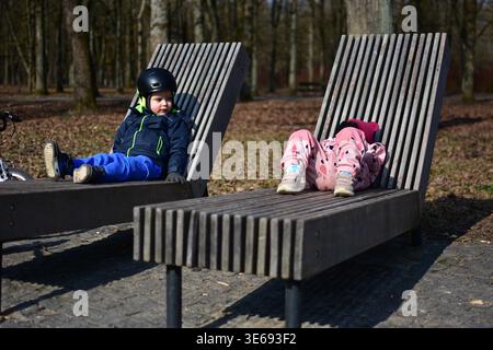 Zwei Kinder entspannen sich an einem sonnigen Frühlingstag auf hölzernen Parkliegen und genießen die Erholung und Freizeit im Freien in einer natürlichen Waldlandschaft. Ausgaben für gleichrangige Elemente Stockfoto
