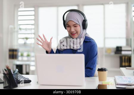 Fröhliche asiatische muslimische Frau, die Videokonferenz auf Laptop in heller Home-Office-Einrichtung hat. Aktivierung virtueller Verbindungen. Stockfoto