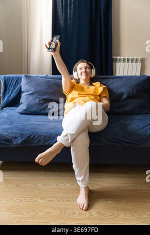 Frau, die Kopfhörer trägt, entspannt sich zu Hause auf einem blauen Sofa und macht fröhlich ein Selfie mit ihrem Smartphone Stockfoto