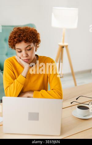 Verschlafene traurige Afroamerikanerin, die mit Laptop am Tisch sitzt. Müde überarbeitetes Mädchen faul zur Arbeit. Geschäftsstudentin, die sich nach der Arbeit am Laptop im Home Office langweilig erholt hat Stockfoto