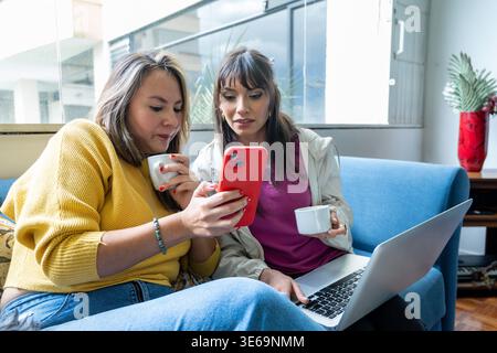 Zwei junge Freundinnen, die auf einer Couch sitzen und Bildschirminhalte auf einem Smartphone teilen Stockfoto