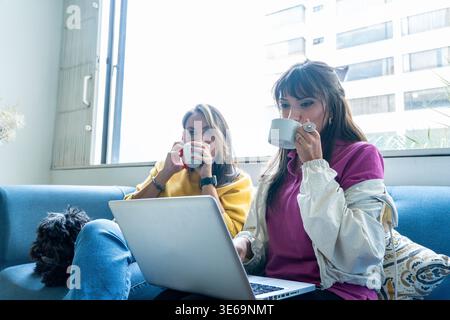 Zwei Freundinnen arbeiten ferngesteuert an einem Laptop und trinken Kaffee in der Wohnung Stockfoto