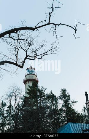 Ein Leuchtturm erhebt sich über der Baumgrenze, während Äste in der Abenddämmerung zum Himmel reichen. Die Struktur ist gegen das verblassende Licht sichtbar, umgeben von Stockfoto