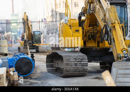 Schwerer gelber Bagger auf einer städtischen Infrastrukturbaustelle mit blauen Wasserrohren auf dem Boden und Stadtgebäuden im Hintergrund während ut Stockfoto