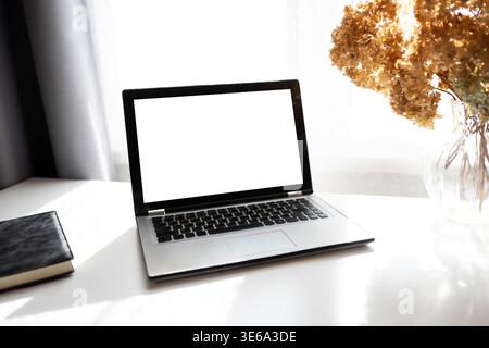 Minimaler Arbeitsbereich mit Laptop auf weißem Schreibtisch in der Nähe von Fensterlicht und getrockneten Blumen. Saubere Geschäftsumgebung für Fernarbeit, Freiberufler und Produktivität Stockfoto
