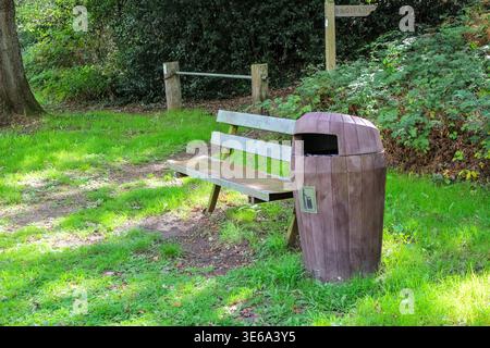 Eine friedliche Parkszene mit einer Holzbank und einem braunen Abfalleimer auf einem üppig grünen Rasen, mit einem Fußwegsschild, das auf einen grünen Weg zeigt. Stockfoto