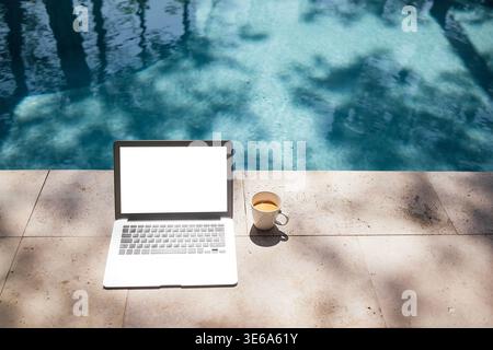 Ein Laptop mit leerem Bildschirm befindet sich auf einer Steinoberfläche neben einem Swimmingpool, mit einer Tasse Kaffee in der Nähe und Bäumen auf dem Wasser Stockfoto