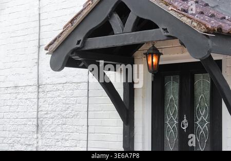 Ein elegantes schwarzes Holzdach mit einer leuchtenden Laterne hängt über einer dunklen Tür mit Glaspaneelen an einer weißen Ziegelwand. Stockfoto