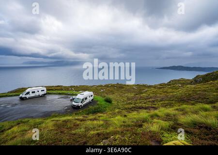 Zwei weiße Wohnmobile parken an einem malerischen Aussichtspunkt in Kråkenes, Norwegen. Die Küstenlandschaft bietet feuchtes grünes Moorland mit Blick auf den dunklen Ozean, Stockfoto