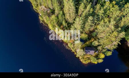 Ein atemberaubendes Luftbild von oben zeigt eine kleine Holzhütte in einem dichten Kiefernwald am Ufer des Flusses Numedalslågen. Das tiefe dunkle Wasser Stockfoto