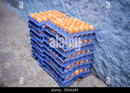 Stapel blauer Eierkartons mit braunen Eiern, die an einer blauen Wand auf einer kopfsteingepflasterten Straße auf einem Markt im Freien aufgestellt sind Stockfoto