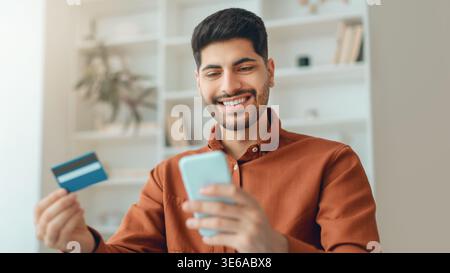 Mann, der das Telefon benutzt, um online einzukaufen, mit Kreditkarte in der Hand in einem hellen Raum Stockfoto