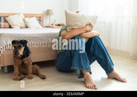 Traurige Frau, die das Gesicht mit einem Kissen bedeckt, Depressionen, Stress und Angst im Schlafzimmer erlebt, mit einem besorgten Hund Stockfoto